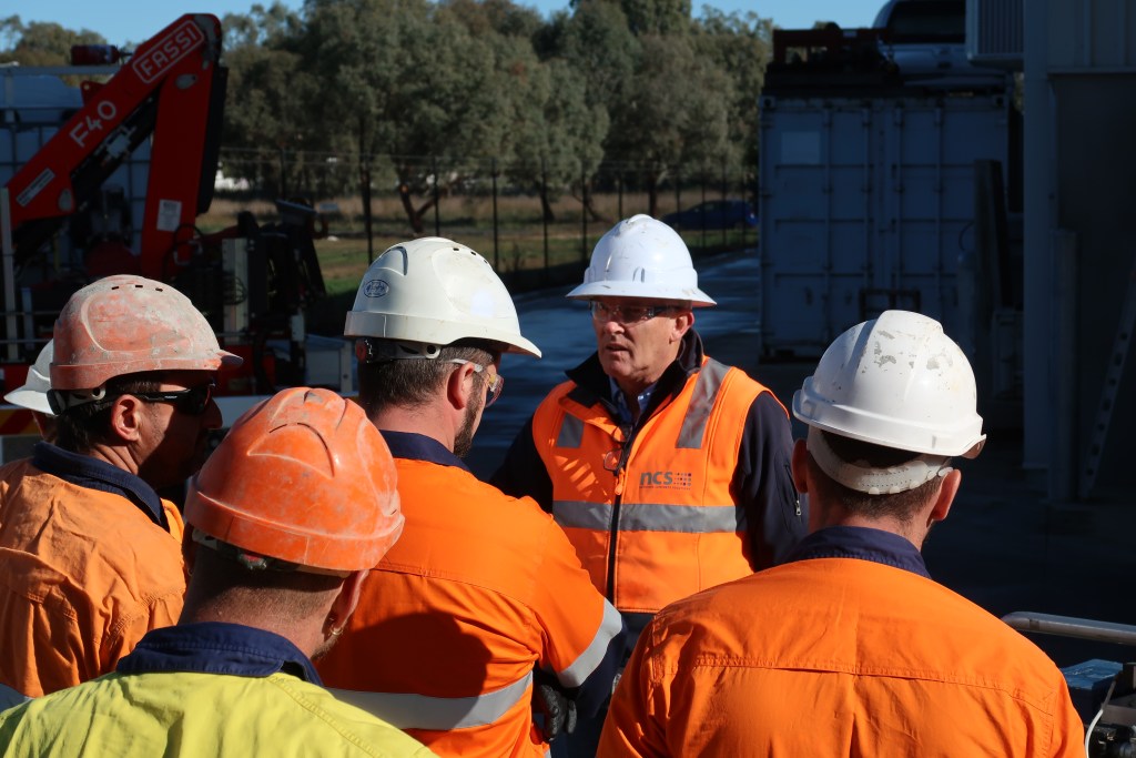 A group of construction workers in high-visibility orange vests and hard hats, gathered around a supervisor who is giving instructions on a worksite.
